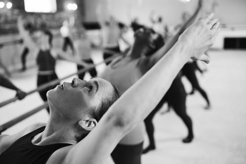 Individual reaching upward with arms extended during a ballet barre class, focusing on balance, flexibility, and full-body engagement in a group fitness setting.