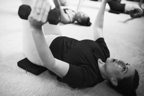 Individual performing a dumbbell chest fly exercise while lying on the floor during a group fitness class, focusing on training the upper body and burning calories.