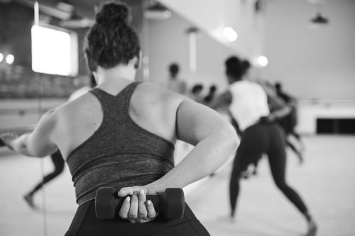 Person in a tank top holding onto the barre with their left hand, arm outstretched, and their right hand holding a small dumbbell with their elbow bent.