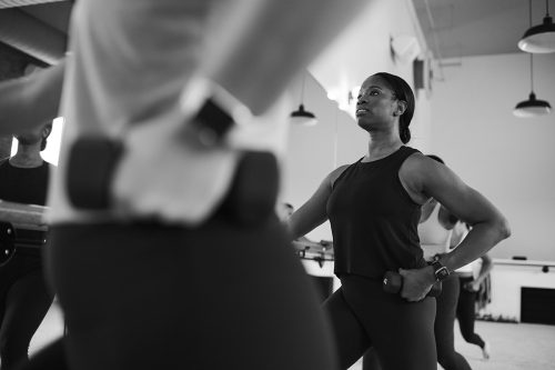 Woman in a tank top holding onto the barre with her right arm outstretched and her left arm bent with hand holding a small dumbbell 