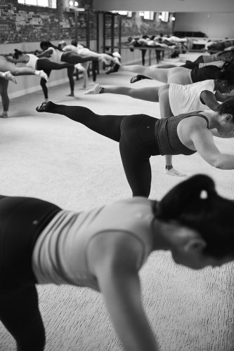 More than a dozen barre students holding onto the barre and backs parallel to floor and their left legs raised with toes pointed out. 