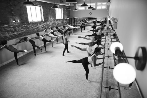 Full-studio view of an instructor teaching several dozen students who are at the barre, holding on with both hands and left legs raised behind them. 