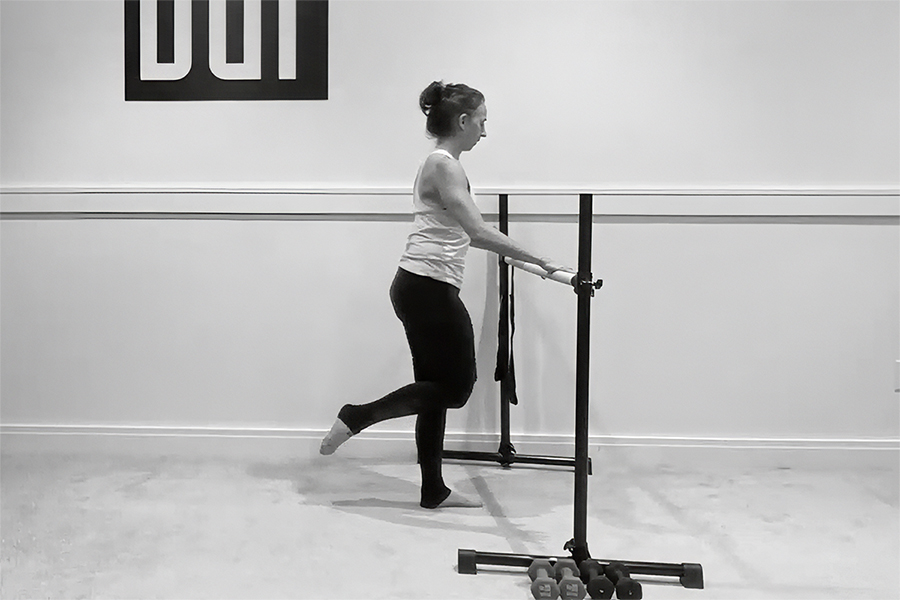 Woman in a barre studio demonstrating a standing seat exercise at the barre.