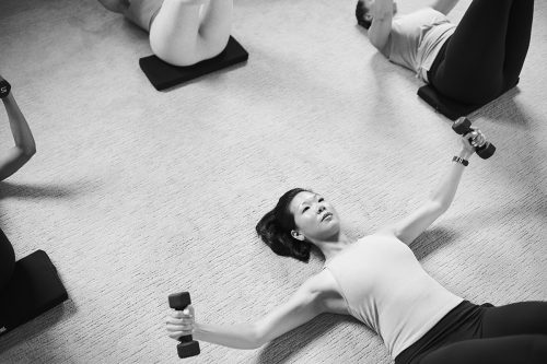 Individual lying on the floor performing a dumbbell chest fly exercise during a serratus anterior workout, focusing on upper body strength in a group fitness class. 