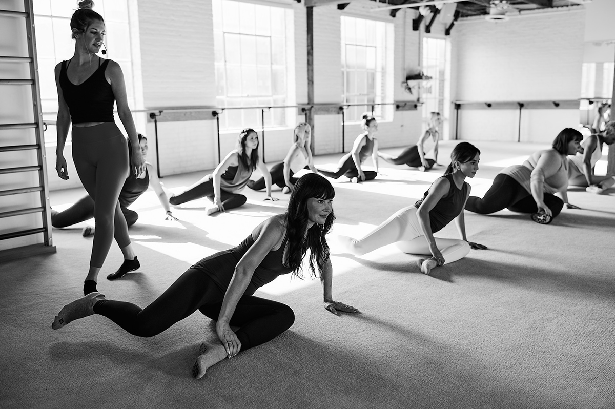 Group of women stretching on the floor in a barre fitness class, guided by an instructor in a spacious, bright studio.