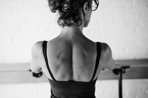 The toned back of a person in a tank top at the barre standing up in a barre class.

