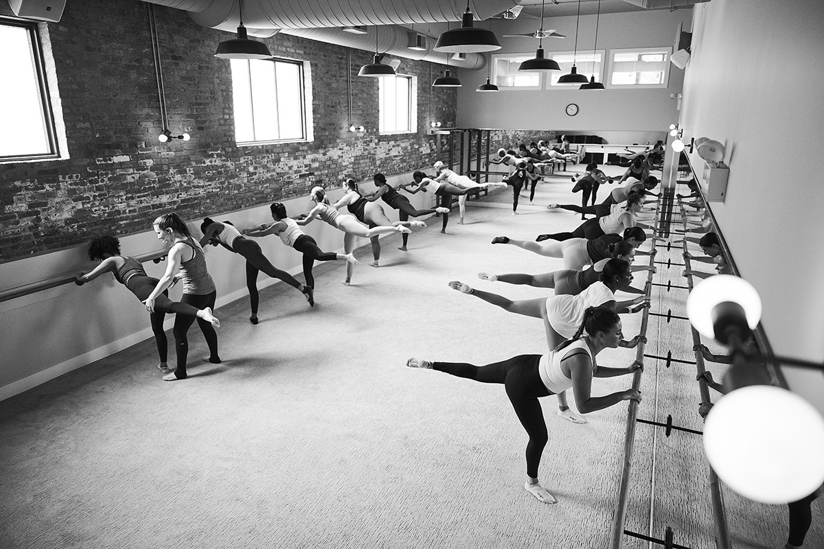 Panoramic of a studio full of students doing leg lifts at the barre during a class. An instructor is helping guide one student with the movement.