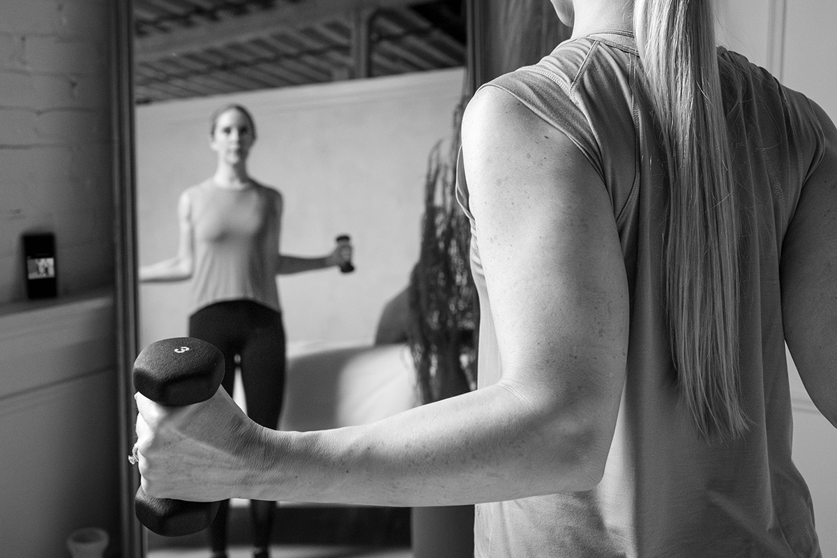A woman is practicing a barre exercise in a mirror using dumbbells.