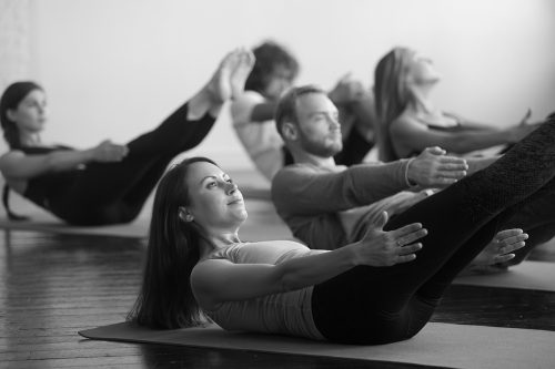 Five students practicing heel taps during barre class, with legs raised together, toes pointed straight, arms tight to sides, and upper back, shoulders and head raised off the ground.