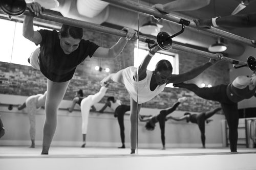 Group of individuals in a barre studio with mirrors running along the walls doing leg lifts at a ballet barre in a fitness class, focusing on balance, performance, flexibility, and core strength. 