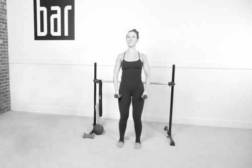 Woman in a Bar Method studio demonstrating shoulder walks with light dumbbells, standing near the ballet barre.