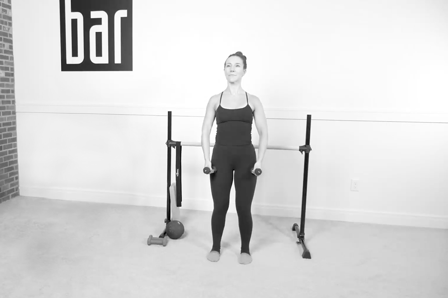 Woman in a Bar Method studio demonstrating shoulder walks with light dumbbells, standing near the ballet barre.