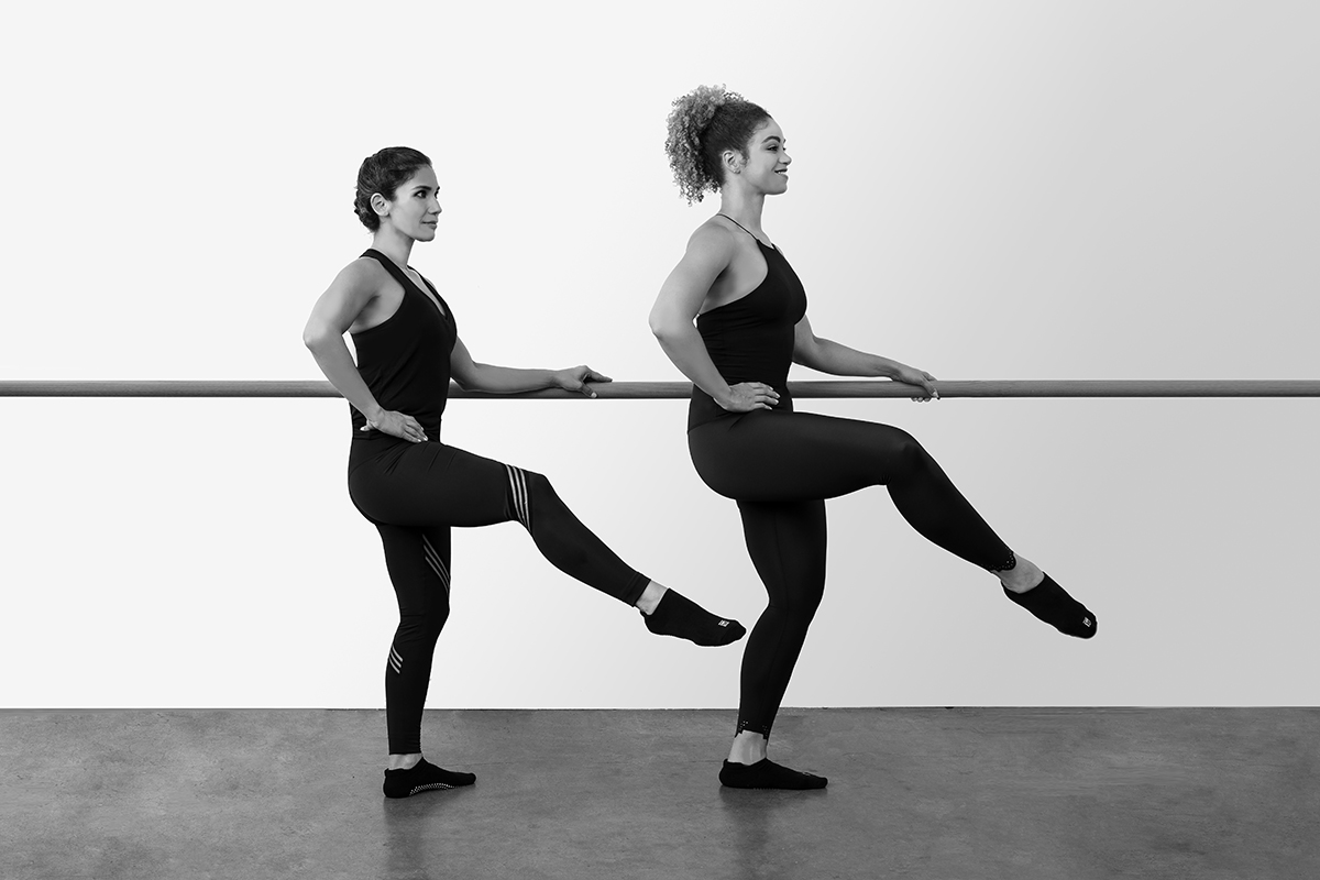 Two women performing leg lifts at the barre in a studio.