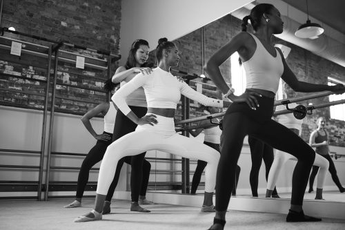 Several women are working out in a barre studio while an instructor provides support and assistance with alignment and form.