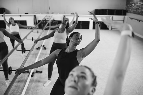 Diverse group of women practicing ballet-inspired moves at the barre, enhancing flexibility and posture in a fitness studio.