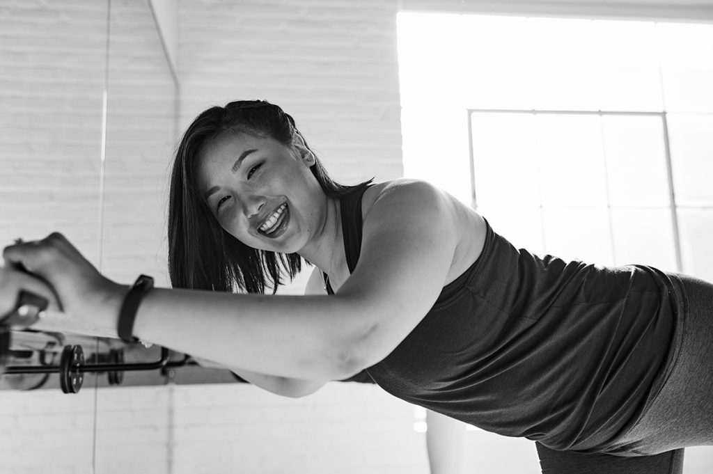 A woman is shown holding onto a bar with both hands and smiling. Her left leg is extended behind her. The room has a large mirror and a white brick wall.