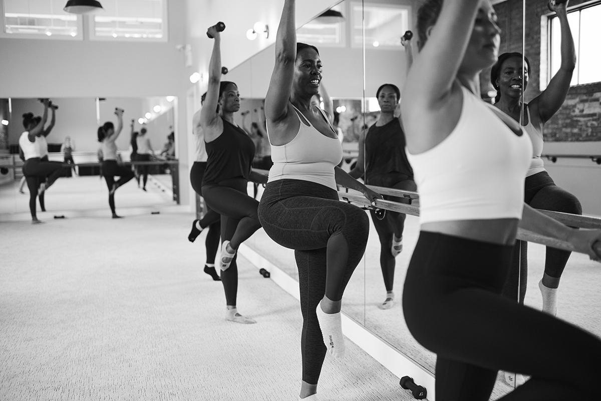 Bar Method students are taking a class. Each student is holding onto the bar with their left hand while lifting their right leg off the ground and reaching their right arm overhead while holding a weight.