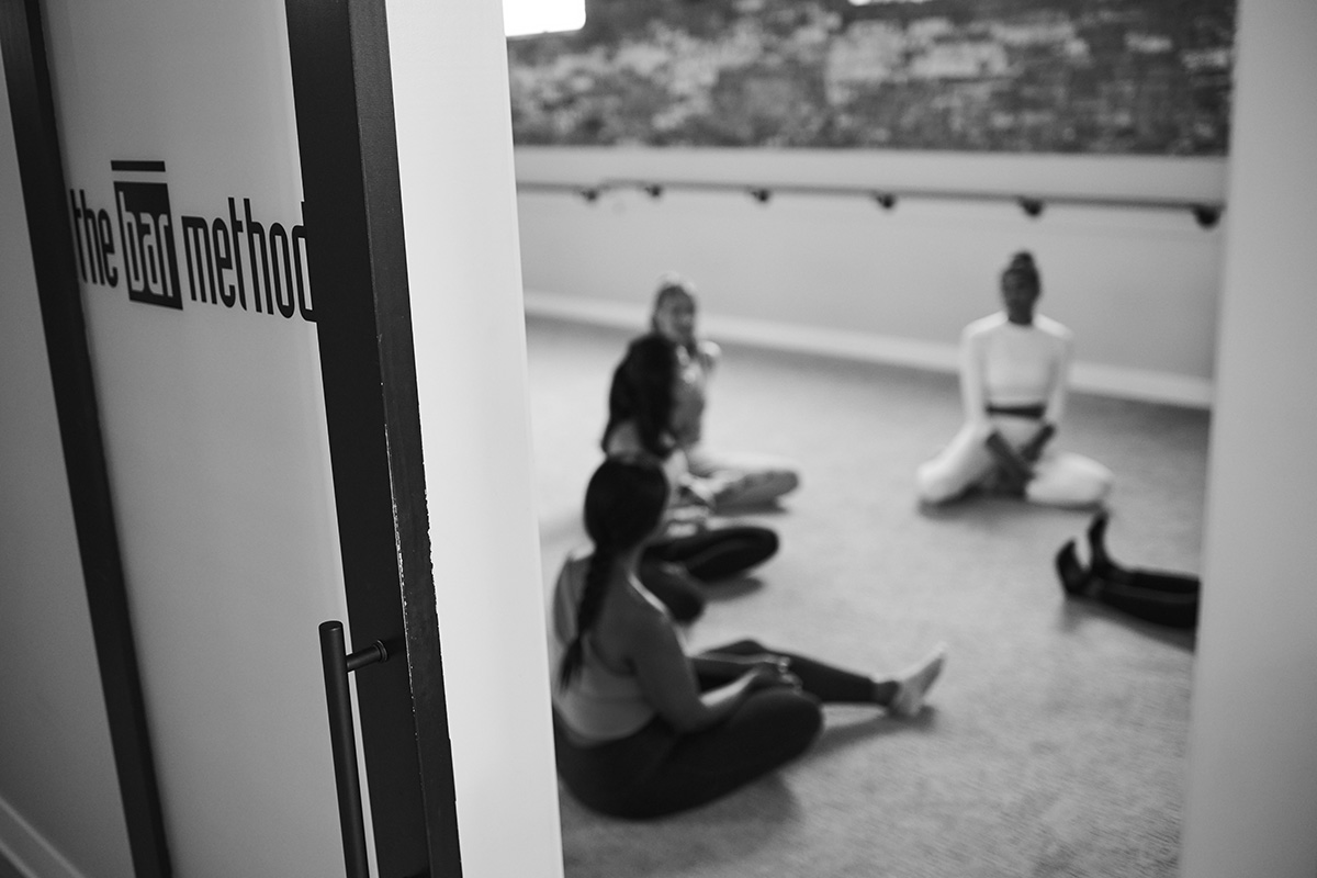 A group of Bar Method members are sitting on the ground in a circle in the studio as part of the Bar Method community.