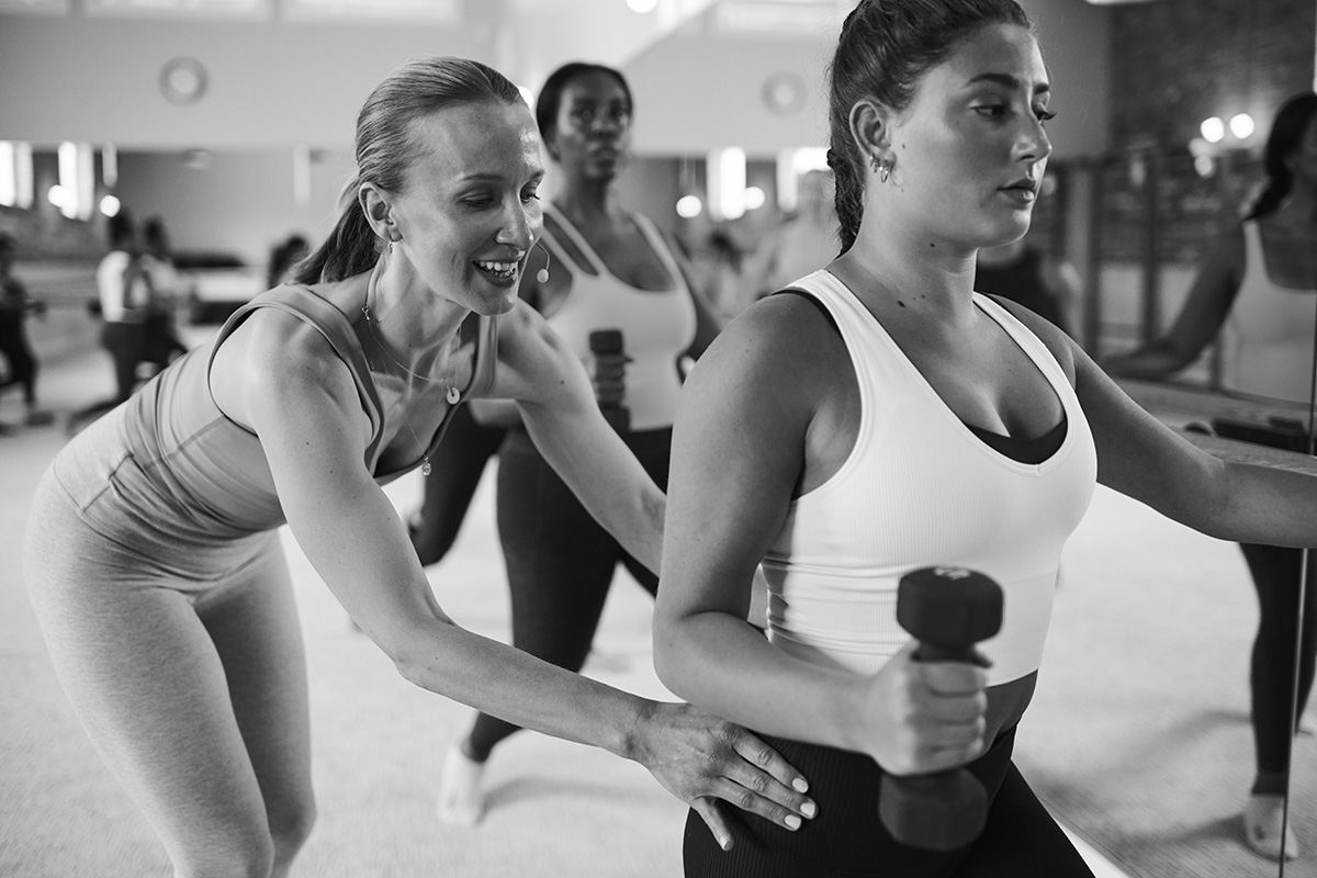 Fitness instructor guiding a woman through a core-strengthening exercise with a dumbbell in a studio class.